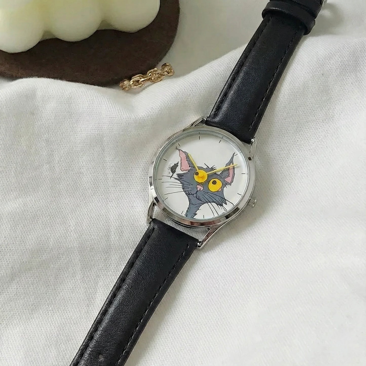 Wristwatch with a cat face design on the dial, black leather strap, on a white fabric background.