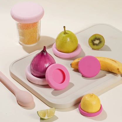 Pink kitchen gadgets on a white surface with fruits and a jar.
